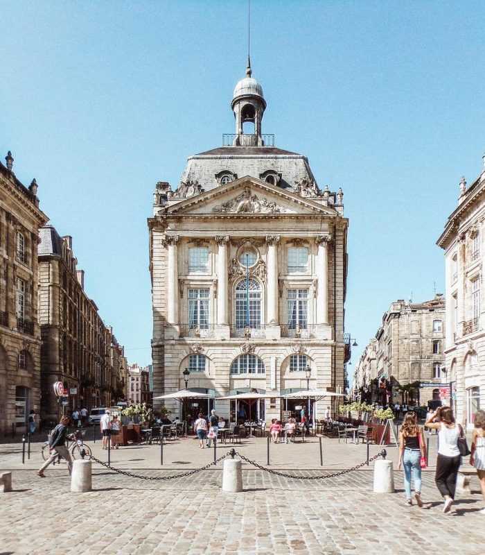 Un grand bâtiment historique avec une horloge et un dôme se dresse sur une place pavée ensoleillée, entourée d'architectures classiques similaires. Les gens marchent, font du vélo et s'assoient à des tables en plein air devant le bâtiment.