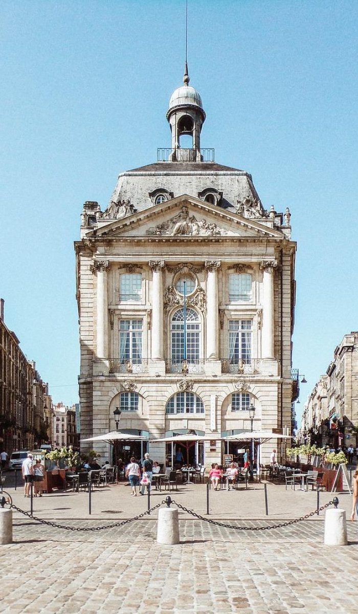 Un grand bâtiment historique avec une horloge et un dôme se dresse sur une place pavée ensoleillée, entourée d'architectures classiques similaires. Les gens marchent, font du vélo et s'assoient à des tables en plein air devant le bâtiment.