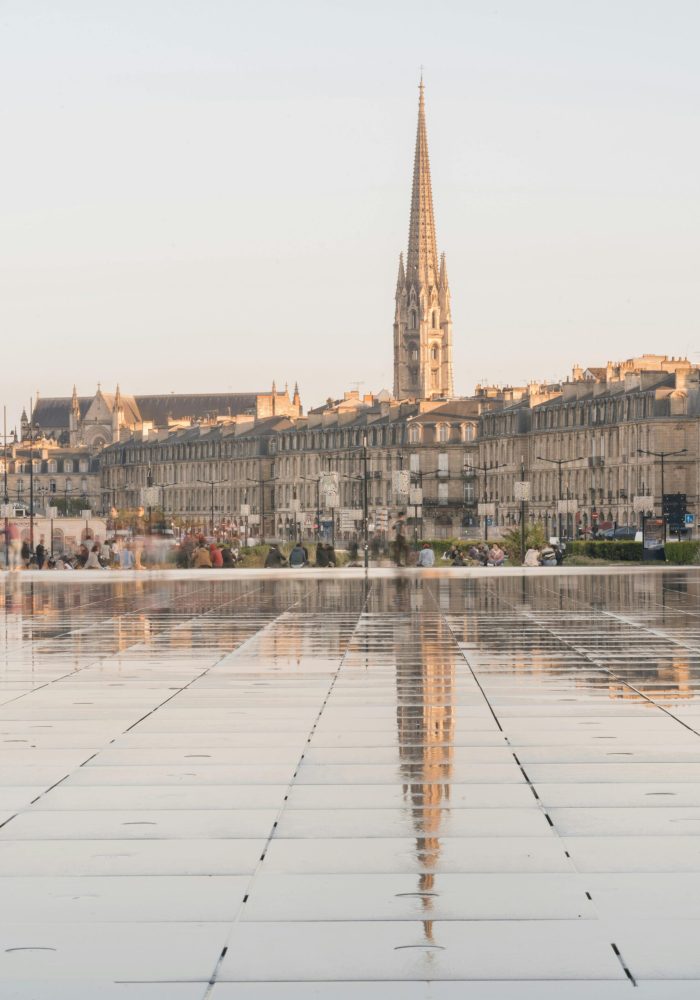 A tall church with a pointed spire is reflected in a large, wet, tiled square, with historic buildings and people in the background in the soft daylight.