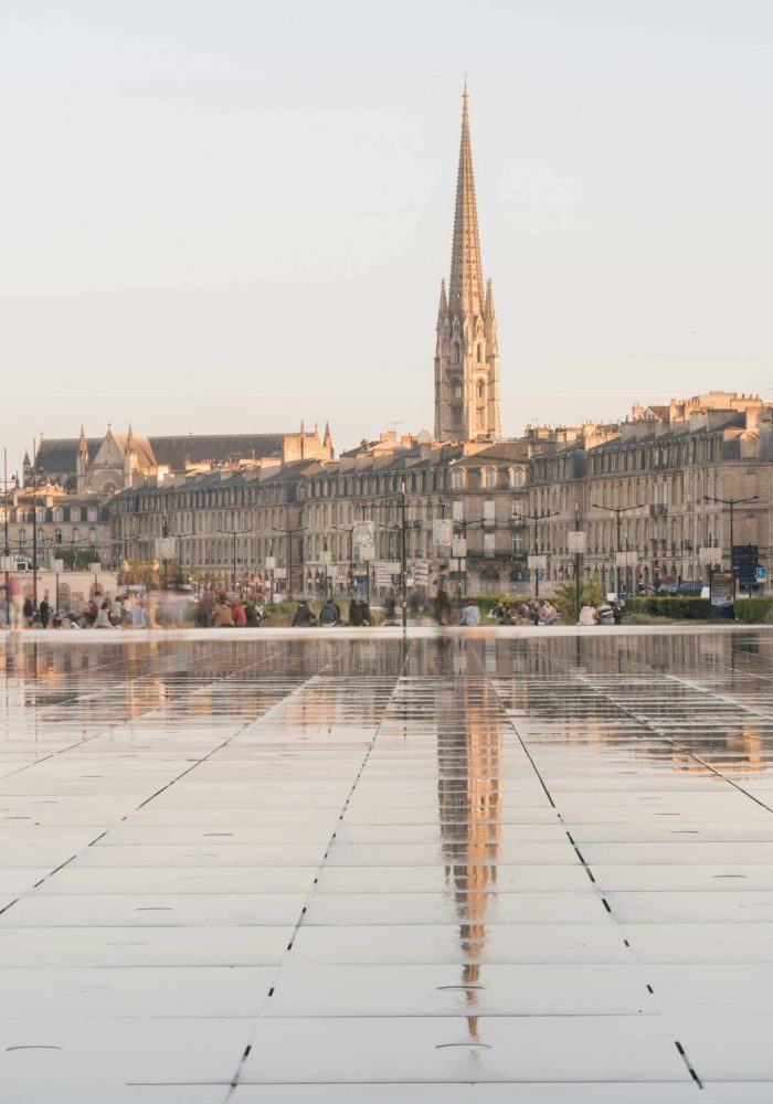 A tall church with a pointed spire is reflected in a large, wet, tiled square, with historic buildings and people in the background in the soft daylight.