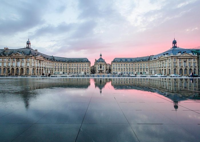 Symmetrical historic buildings reflected in a large shallow mirror of water at sunset, with a pink and blue sky overhead. The scene is peaceful and showcases classic European architecture.