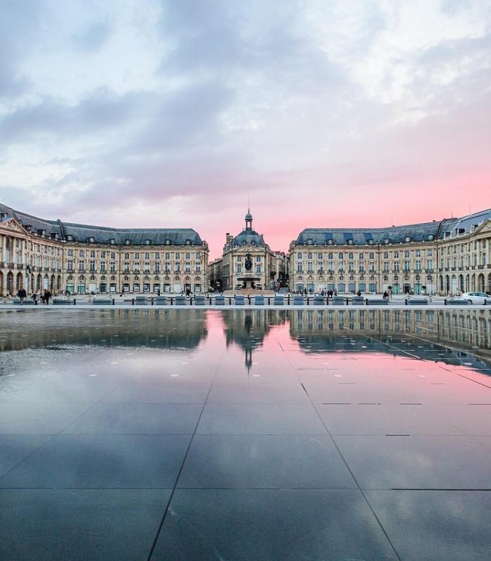 Bâtiments historiques symétriques se reflétant dans un grand miroir d'eau peu profond au coucher du soleil, avec un ciel rose et bleu au-dessus de la tête. La scène est paisible et met en valeur l'architecture européenne classique.