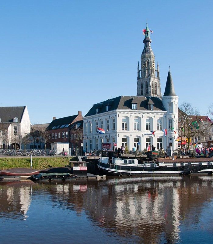 Un bâtiment historique blanc avec une haute tour d'horloge se dresse près d'un canal avec des bateaux amarrés ; un ciel bleu clair et des personnes rassemblées autour du bâtiment sont visibles.