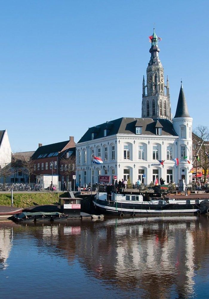 Un bâtiment historique blanc avec une haute tour d'horloge se dresse près d'un canal avec des bateaux amarrés ; un ciel bleu clair et des personnes rassemblées autour du bâtiment sont visibles.