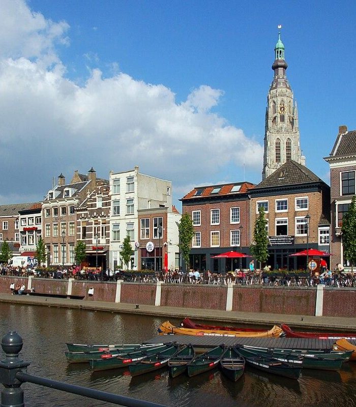 Un canal avec des bateaux amarrés devant des bâtiments historiques et un grand clocher sous un ciel bleu à Breda, Pays-Bas. Des personnes sont assises à des cafés en plein air le long du front de mer.