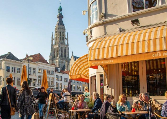 Des personnes sont assises à des tables de café en plein air sous des auvents rayés de jaune, lors d'une journée ensoleillée sur une place d'une ville européenne, avec un grand clocher d'église et des bâtiments historiques en arrière-plan.