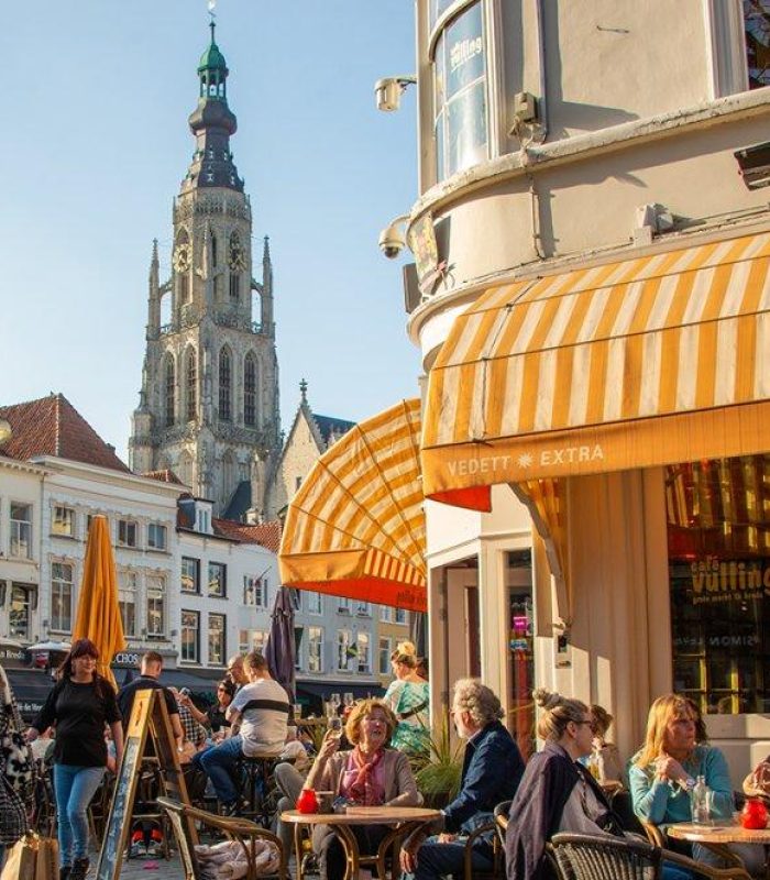 Des personnes sont assises à des tables de café en plein air sous des auvents rayés de jaune, lors d'une journée ensoleillée sur une place d'une ville européenne, avec un grand clocher d'église et des bâtiments historiques en arrière-plan.