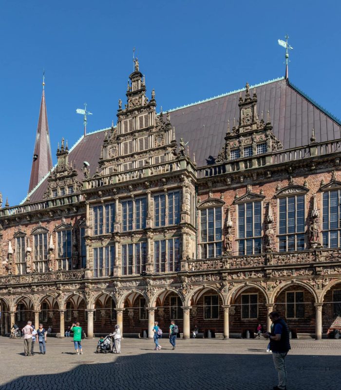 Un bâtiment historique orné de détails architecturaux gothiques et Renaissance se dresse sur une place ensoleillée, avec des gens qui marchent et un ciel bleu au-dessus. Des statues et de grandes fenêtres ornent la façade.