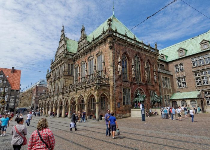 Les gens marchent et se rassemblent sur une place pavée devant un bâtiment historique en briques orné d'arches, de statues et d'un toit en cuivre vert, sous un ciel partiellement nuageux.