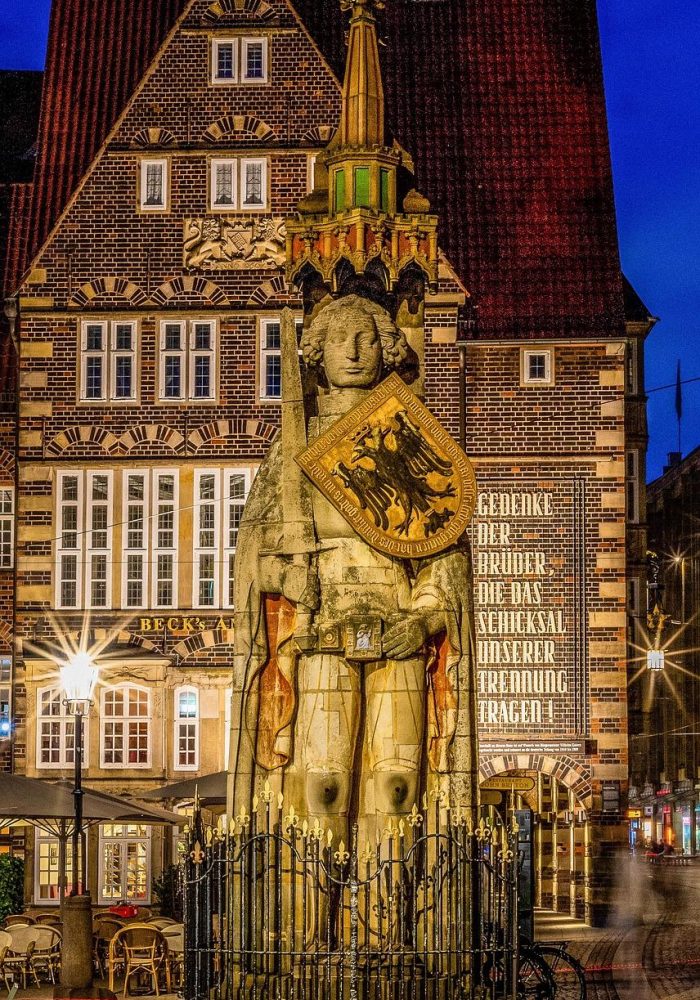 Une grande statue de Roland tenant un bouclier avec un aigle noir se dresse sur une place de la ville, entourée d'une clôture en fer forgé. Derrière elle se trouvent des bâtiments historiques en briques avec des fenêtres éclairées et une enseigne.