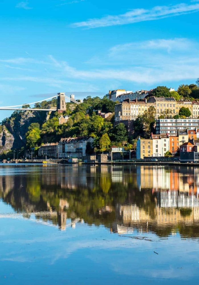 View of the Clifton suspension bridge over the Avon Gorge in Bristol, England, with colourful houses and greenery reflected in the calm river under a bright blue sky.