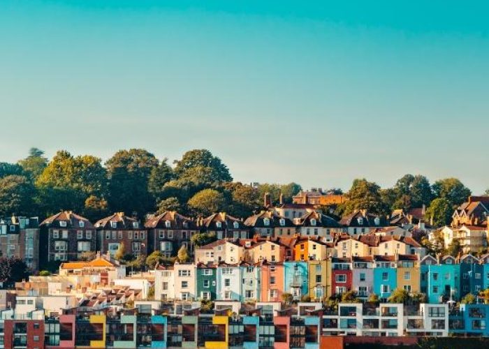 Des maisons et des immeubles colorés se dressent en rangs serrés sur le flanc d'une colline, entourés d'arbres verts sous un ciel bleu limpide.