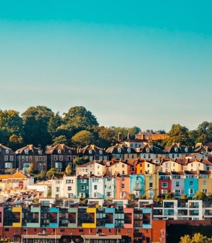 Colourful houses and buildings stand in neat rows on a hillside, surrounded by green trees under a clear blue sky.