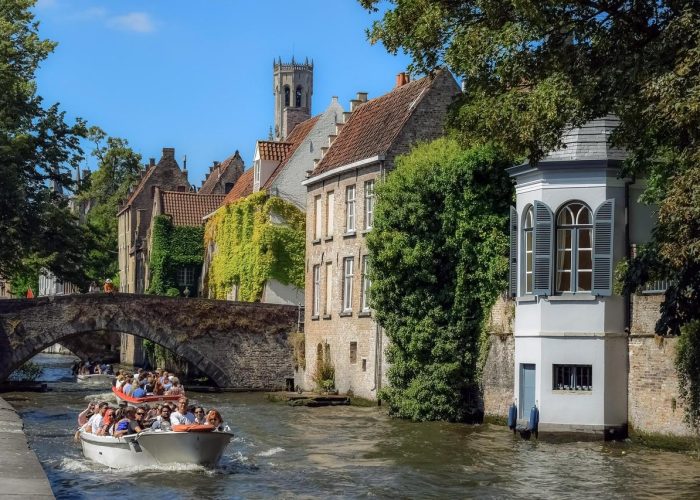 Un bateau rempli de touristes descend un canal bordé de bâtiments historiques en briques et de feuillages verts à Bruges, en Belgique. Un pont de pierre enjambe l'eau et un clocher s'élève à l'arrière-plan sous un ciel bleu.