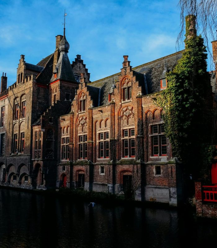 Des bâtiments historiques en brique aux pignons ornés bordent un canal étroit sous un ciel bleu, avec des reflets dans l'eau et de la verdure grimpant le long des murs.