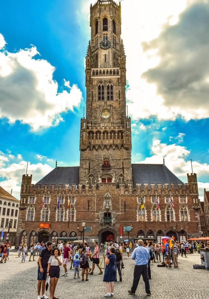 People walk along a busy square in front of Bruges' medieval Belfry, a tall ornate bell tower with a clock, under a partly cloudy blue sky in Bruges, Belgium.