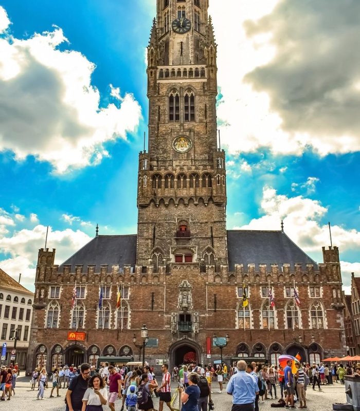 Des personnes marchent sur une place animée devant le Beffroi médiéval de Bruges, un haut clocher orné avec une horloge, sous un ciel bleu partiellement nuageux à Bruges, en Belgique.