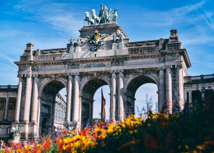 Arc de triomphe avec trois grands arcs et un quadrige en bronze au sommet, sur fond de ciel bleu ; des fleurs colorées occupent le premier plan.