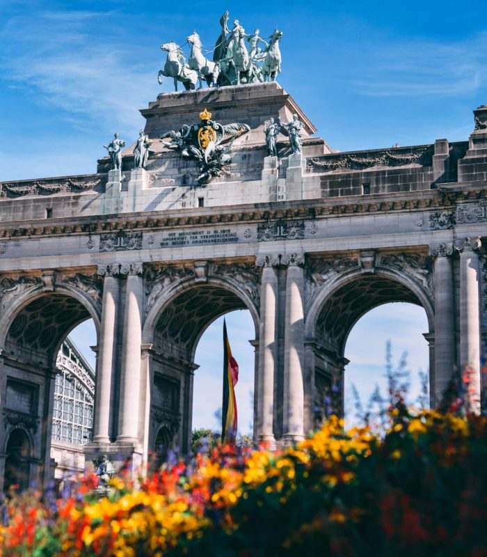 Triumphal arch with three large arches and a bronze quadriga at the top, against a blue sky; colourful flowers in the foreground.