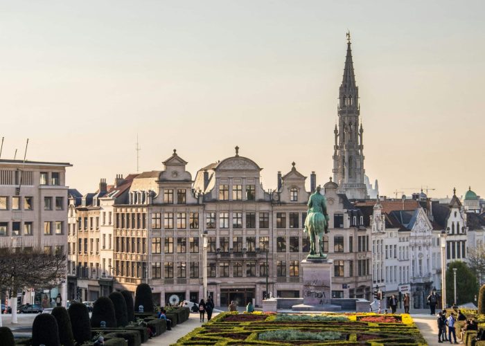 Paysage urbain de Bruxelles vu d'un jardin, avec des bâtiments historiques, la statue d'un cavalier à cheval et la haute flèche de l'hôtel de ville de Bruxelles à l'arrière-plan. Des personnes se promènent et se détendent dans le parc.