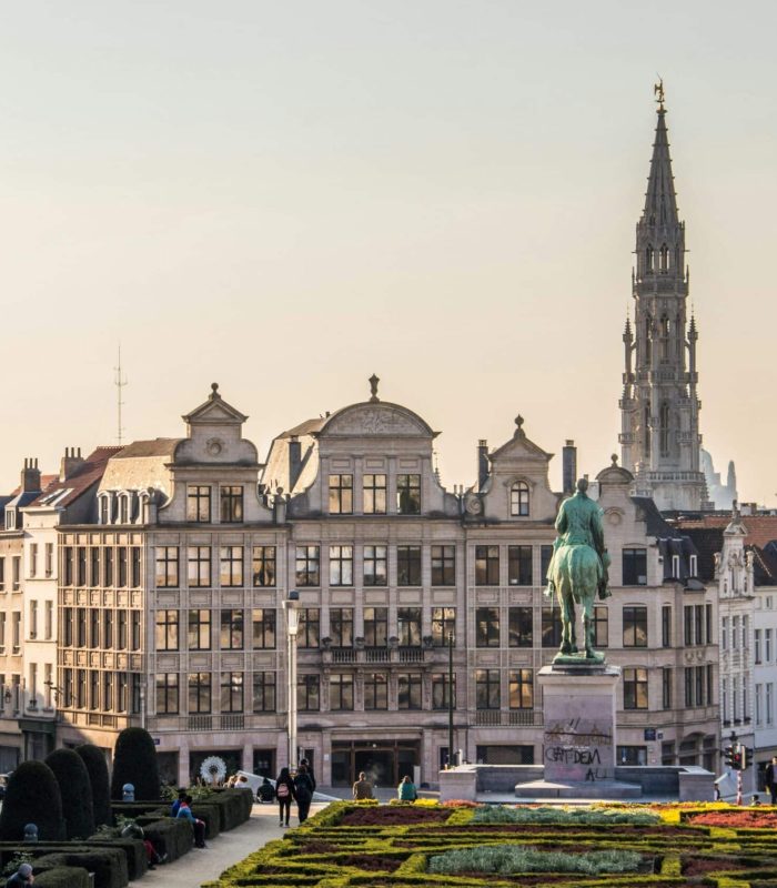 Cityscape of Brussels seen from a garden, with historic buildings, the statue of a rider on horseback and the tall spire of Brussels City Hall in the background. People strolling and relaxing in the park.