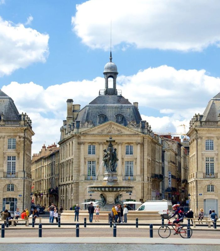 Un grand bâtiment historique avec un dôme se dresse derrière une fontaine et une place, où les gens se promènent et font du vélo sous un ciel bleu. La scène se reflète dans un bassin peu profond, capturant l'atmosphère animée idéale pour le portage bagages événements ou la consigne bagages France services.