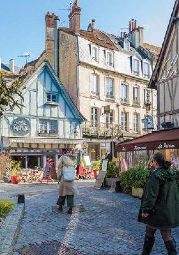 Une charmante scène de rue européenne avec des gens qui marchent, des bâtiments à colombages colorés, des restaurants en plein air et de la verdure par une journée ensoleillée. Des panneaux en français sont visibles sur les bâtiments.