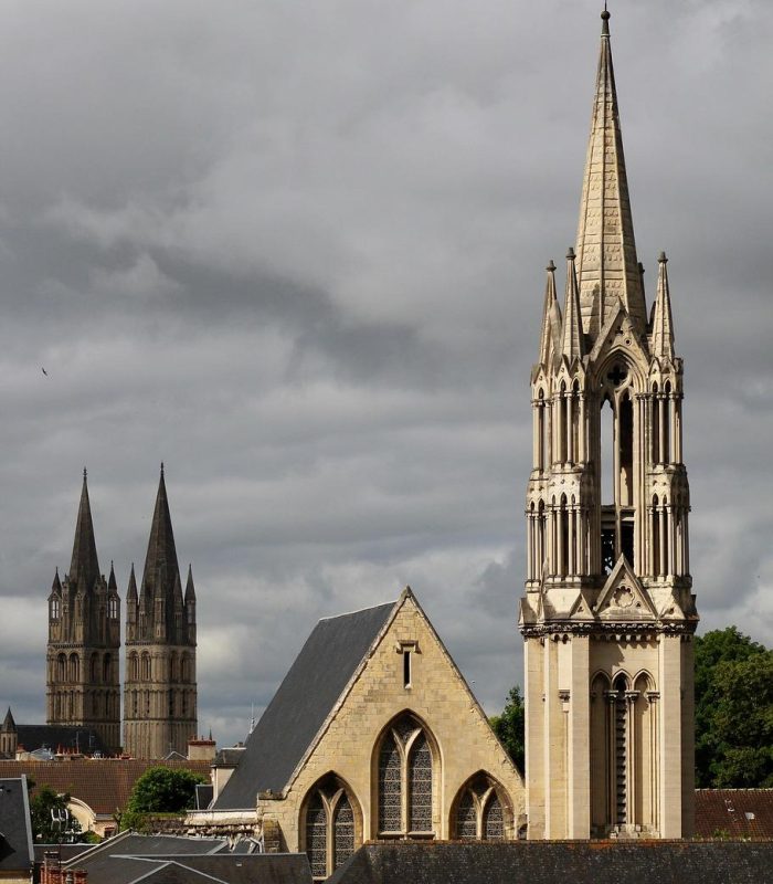 Vue d'une ville avec une église importante dotée d'une haute flèche ornée au premier plan et des flèches jumelles d'un autre bâtiment historique à l'arrière-plan, sous un ciel nuageux.