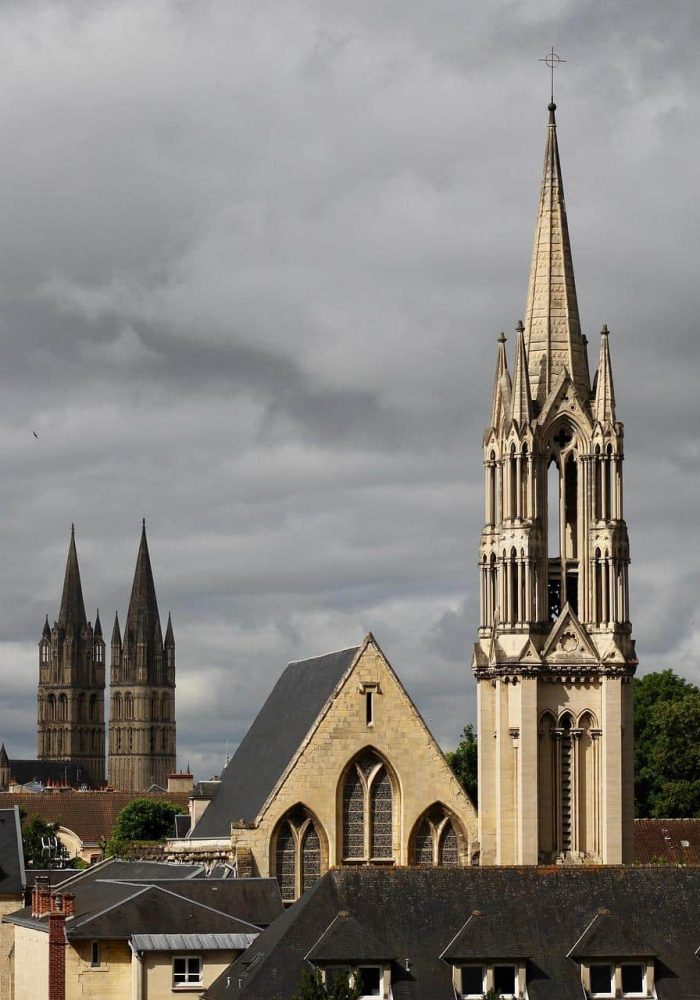 View of a town with a major church with a tall ornate spire in the foreground and the twin spires of another historic building in the background, under a cloudy sky.