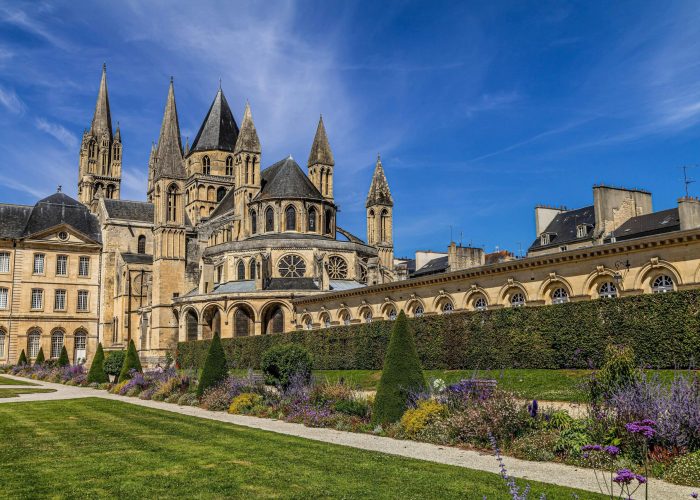 A large historic stone abbey, with tall spires and ornate towers, stands behind a manicured garden, blooming with purple and green plants, under a bright blue sky with billowing clouds.
