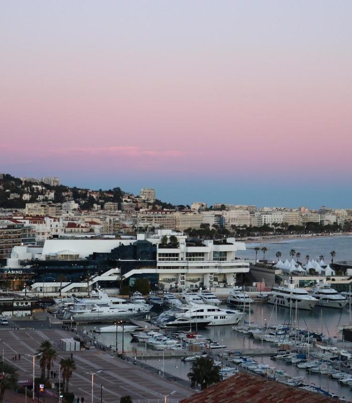 Un port de plaisance rempli de yachts et de voiliers se trouve à côté d'une ville côtière au coucher du soleil. Des bâtiments bordent le rivage et des collines s'élèvent à l'arrière-plan sous un ciel rose et bleu.