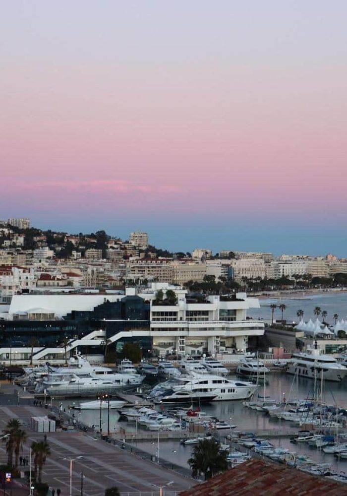 A marina full of yachts and sailboats stands next to a coastal town at sunset. Buildings line the shore and hills rise in the background under a pink and blue sky.