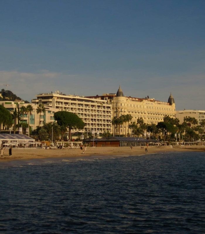 View of a sandy beach in Cannes, France, with people walking along the shore, palm trees and elegant historic buildings lining the seafront, under a clear blue sky.