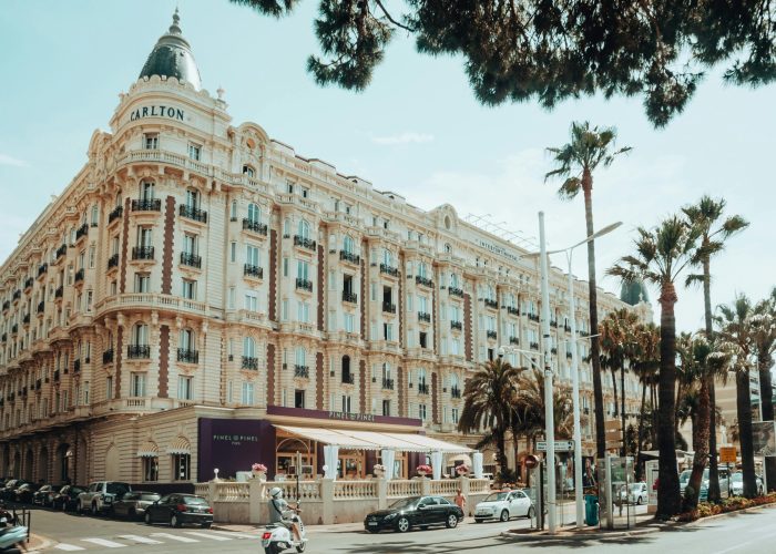 Vue de la rue du grand hôtel Carlton à Cannes, France, avec une architecture ornée, des palmiers, des voitures garées, des scooters, et des personnes dînant dans un café sur le trottoir sous un auvent violet par une journée ensoleillée.