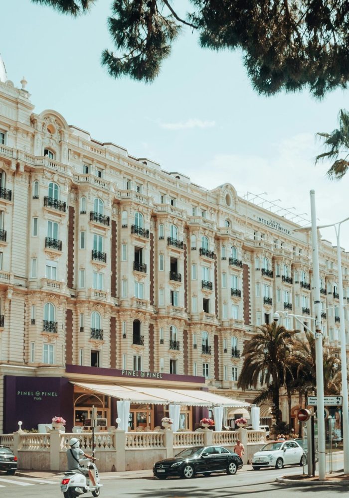 Vue de la rue du grand hôtel Carlton à Cannes, France, avec une architecture ornée, des palmiers, des voitures garées, des scooters, et des personnes dînant dans un café sur le trottoir sous un auvent violet par une journée ensoleillée.