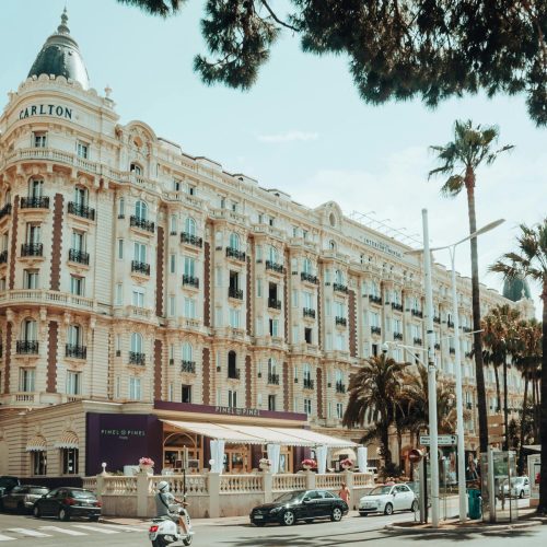 Vue de la rue du grand hôtel Carlton à Cannes, France, avec une architecture ornée, des palmiers, des voitures garées, des scooters, et des personnes dînant dans un café sur le trottoir sous un auvent violet par une journée ensoleillée.