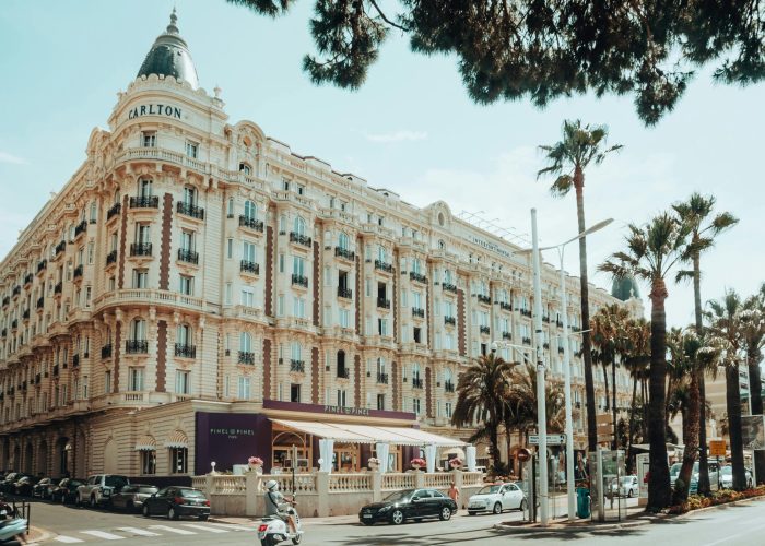 Street view of the Grand Hotel Carlton in Cannes, France, with ornate architecture, palm trees, parked cars, scooters, and people dining in a café on the pavement under a purple awning on a sunny day.