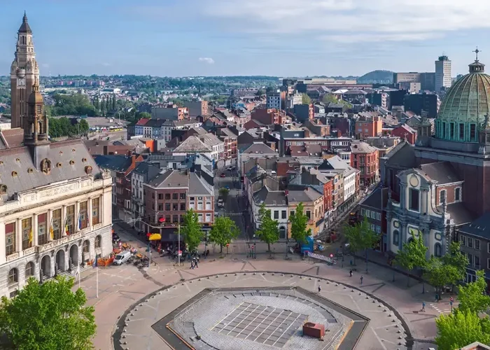 Vue aérienne d'une place de ville européenne avec des bâtiments historiques, dont une église à coupole et une tour d'horloge, entourée d'arbres, de personnes et d'un mélange d'architecture ancienne et moderne sous un ciel partiellement nuageux.