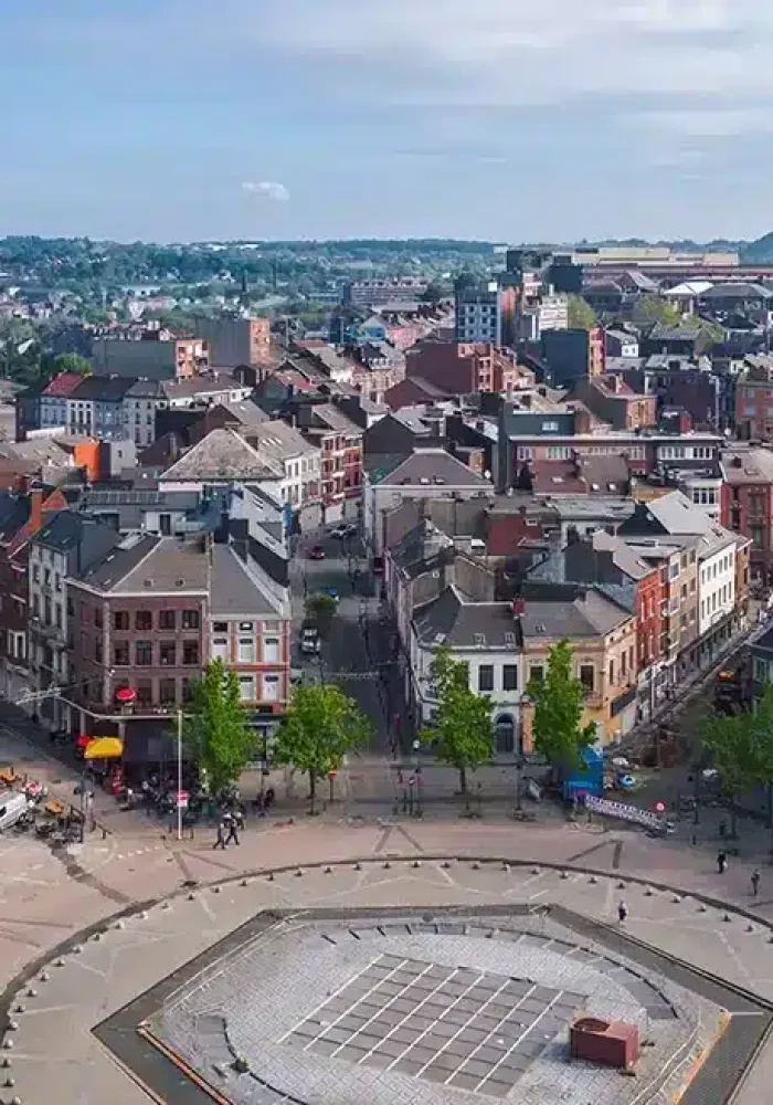 Vue aérienne d'une place de ville européenne avec des bâtiments historiques, dont une église à coupole et une tour d'horloge, entourée d'arbres, de personnes et d'un mélange d'architecture ancienne et moderne sous un ciel partiellement nuageux.