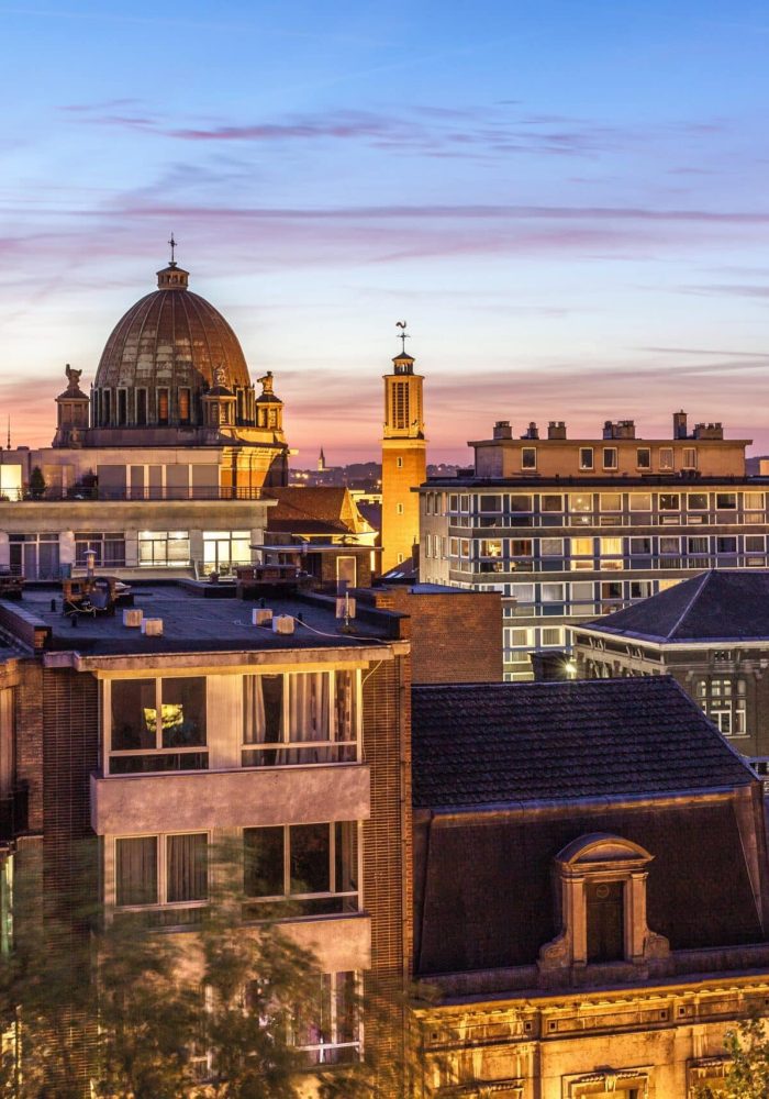 Un paysage urbain au coucher du soleil avec des bâtiments historiques, une structure en dôme, une tour d'horloge et des toits modernes, le tout sous un ciel coloré et crépusculaire avec de doux nuages.