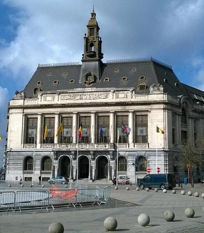 Un grand bâtiment historique avec des drapeaux au-dessus de son entrée se dresse sur une place de la ville, entouré de bornes et de grilles. Une grande tour d'horloge se dresse à l'arrière-plan, sous un ciel partiellement nuageux.