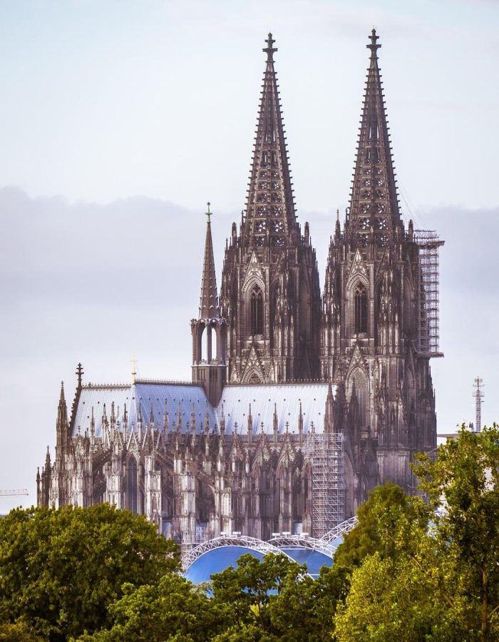 Une grande cathédrale gothique dotée de deux hautes flèches ornées s'élève au-dessus d'arbres verts sous un ciel nuageux. Des échafaudages sont visibles sur une partie de la structure.