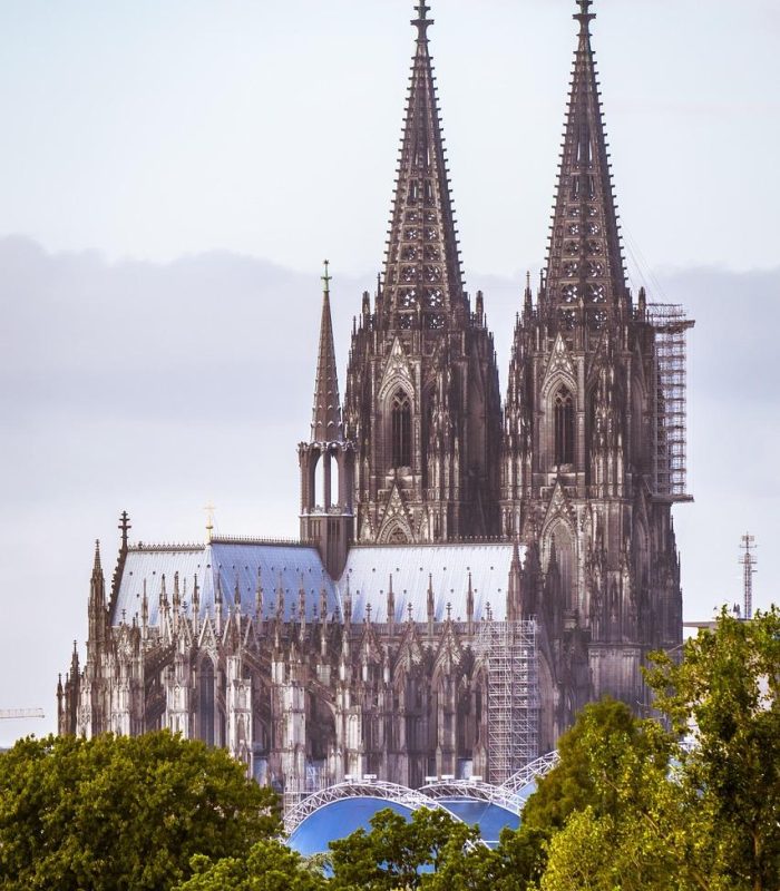A large Gothic cathedral with two tall, ornate spires rises above green trees under a cloudy sky. Scaffolding is visible on part of the structure.