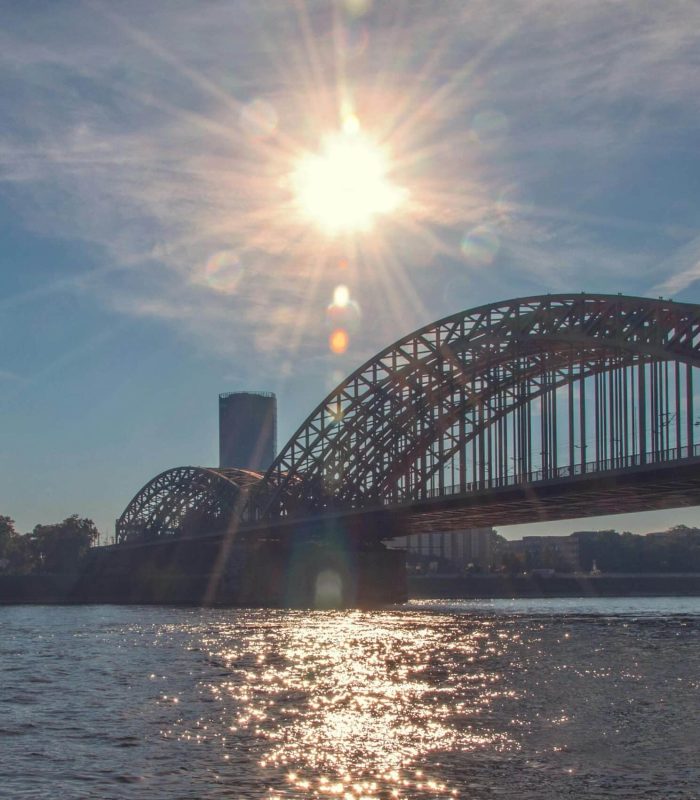 Le soleil brille au-dessus d'un grand pont arqué qui enjambe une large rivière, avec des reflets scintillants sur l'eau et quelques bâtiments visibles à l'arrière-plan sous un ciel bleu clair.