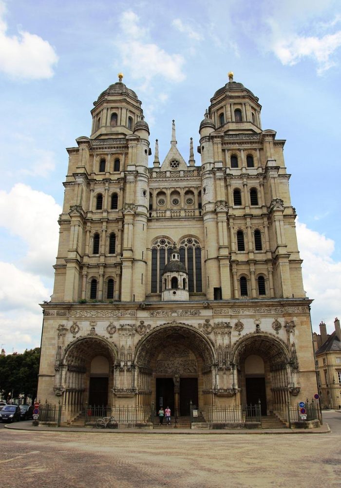 Under a partly cloudy sky stands a large stone church adorned with two high towers and detailed arches. The façade features columns, decorative carvings and three main doors. A few people can be seen near the entrance.