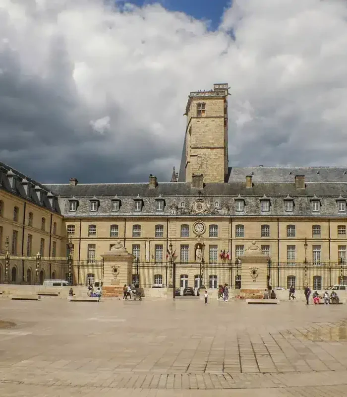 A large historic building with a central tower and symmetrical wings, set around a vast cobbled square. People stroll around and sit at tables in the open air under a partly cloudy sky.