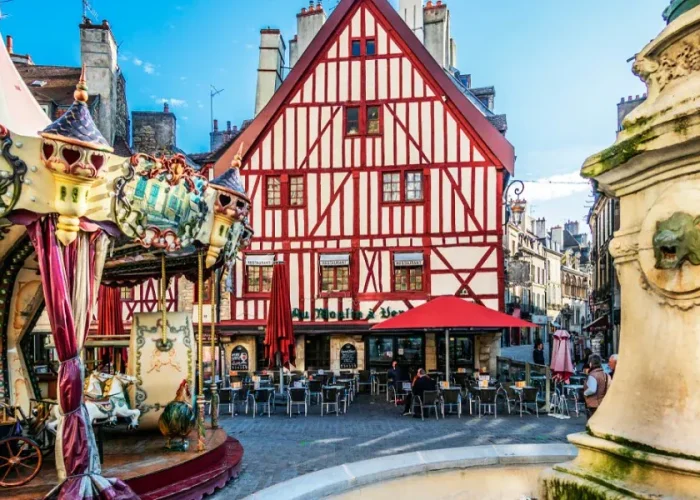 Un bâtiment à colombages coloré avec des rayures rouges et blanches se dresse derrière un café en plein air, à côté d'un manège et d'une fontaine en pierre sur une place européenne animée sous un ciel bleu.