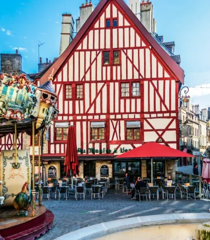 Un bâtiment à colombages coloré avec des rayures rouges et blanches se dresse derrière un café en plein air, à côté d'un manège et d'une fontaine en pierre sur une place européenne animée sous un ciel bleu.