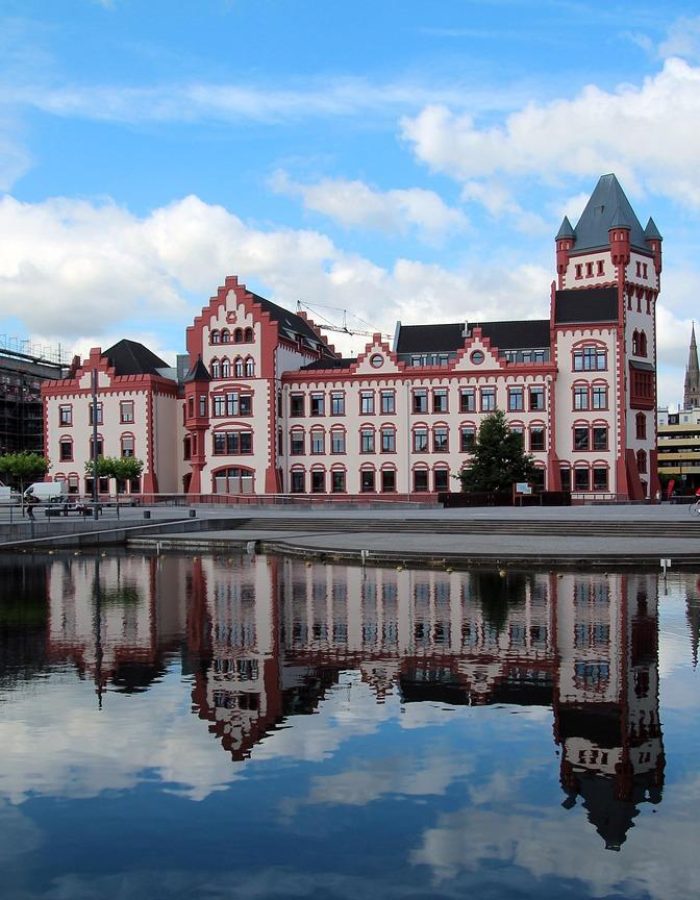 Un grand bâtiment orné de briques rouges et blanches se reflète dans une étendue d'eau calme. Le ciel est partiellement nuageux, il y a des arbres et des gens qui se promènent à proximité. D'autres bâtiments sont visibles à l'arrière-plan.
