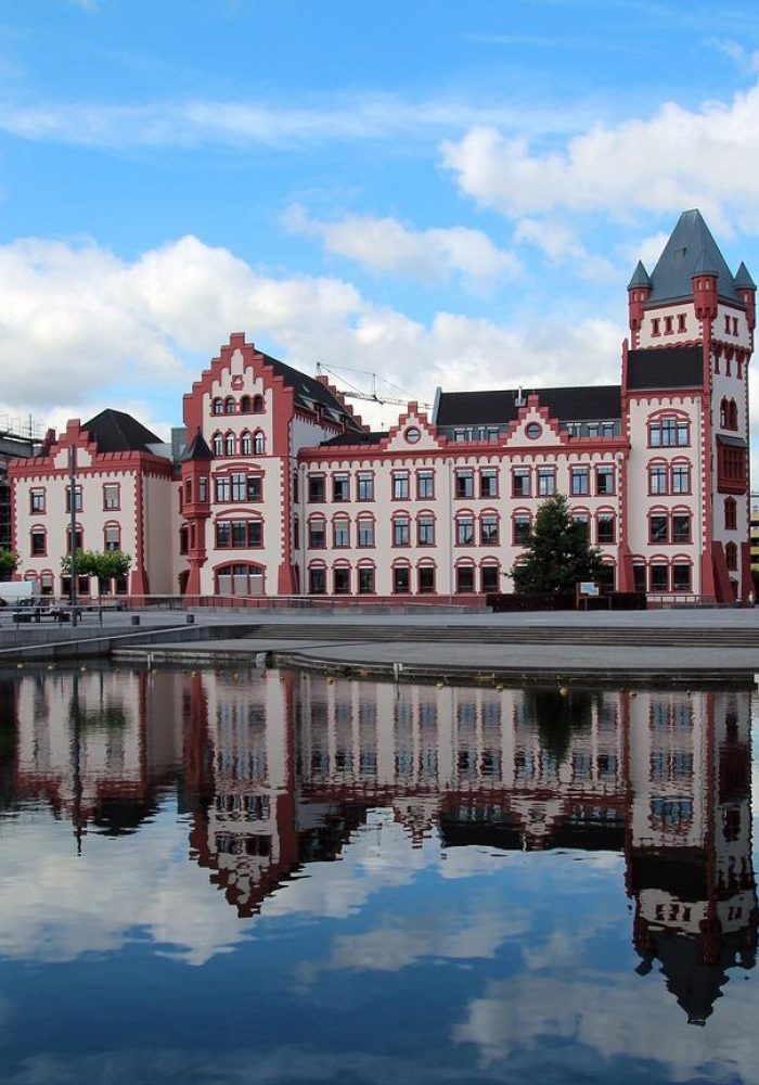 A large building decorated with red and white bricks is reflected in a calm stretch of water. The sky is partly cloudy, and there are trees and people walking nearby. Other buildings can be seen in the background.